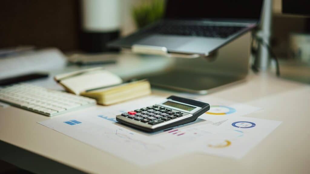 Calculator on a desk next to printed charts, a keyboard and a laptop